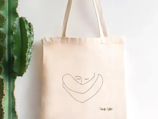 Tote-bag Confiance en soi accroché à un porte-manteau, avec un tracé représentant une personne qui se prend elle-même dans ses bras, et le logo Pochi Kibo en bas à droite, fond blanc avec un cactus à gauche.