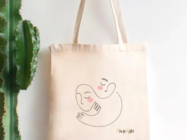 Tote-bag Réconfort beige avec un tracé représentant deux personnes qui s’enlacent, chacune avec une joue rose avec le logo Pochi Kibo en bas à droite, accroché à un porte-manteau et un cactus à gauche sur fond blanc.